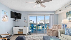 A spacious living room with a large window, a ceiling fan, a flat-screen TV, and a comfortable seating area.