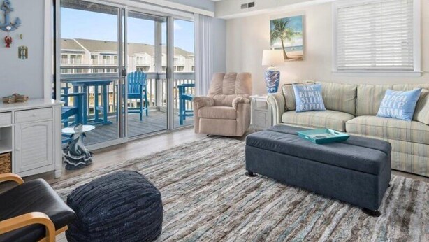A spacious living room with a large sliding glass door leading to a balcony, a beige sofa, a dark ottoman, and a wooden coffee table.
