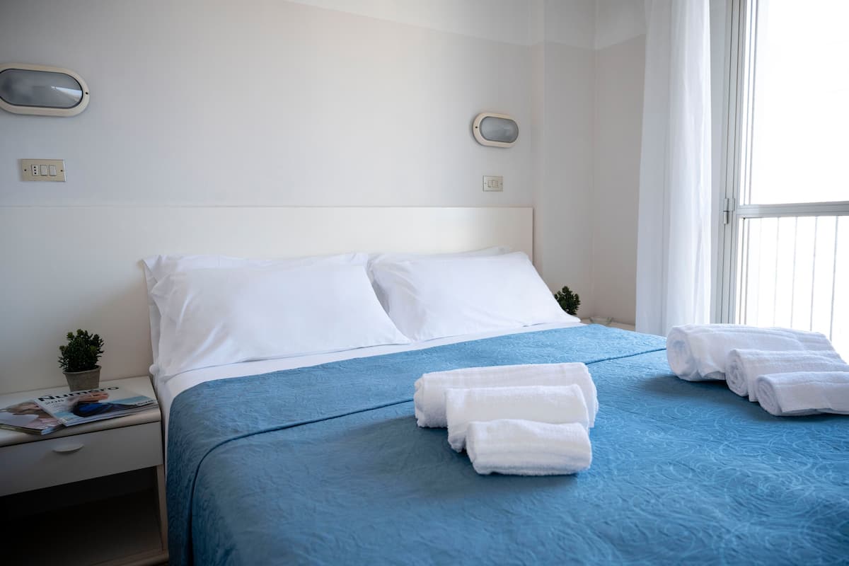 A hotel room with a bed, white pillows, a blue bedspread, and rolled white towels on the bed.