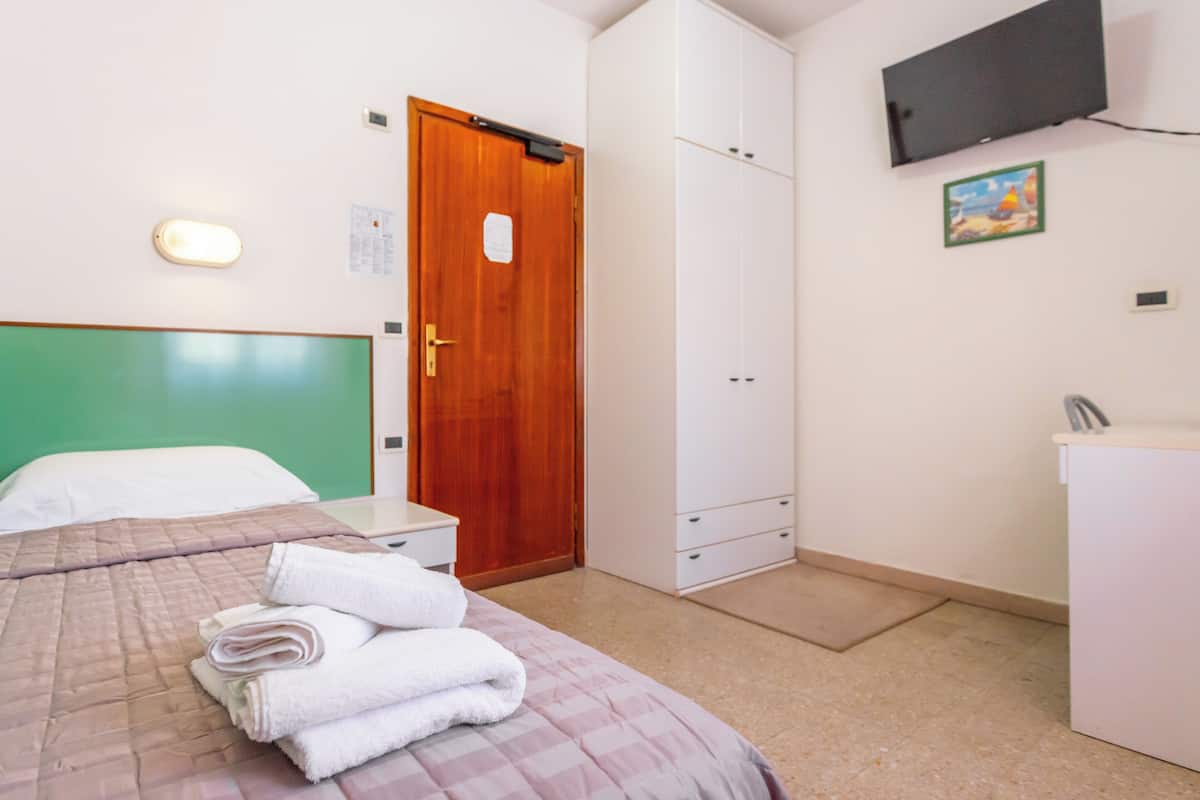 A hotel room with a bed, a wooden door, a white wardrobe, a small desk, and a television mounted on the wall.