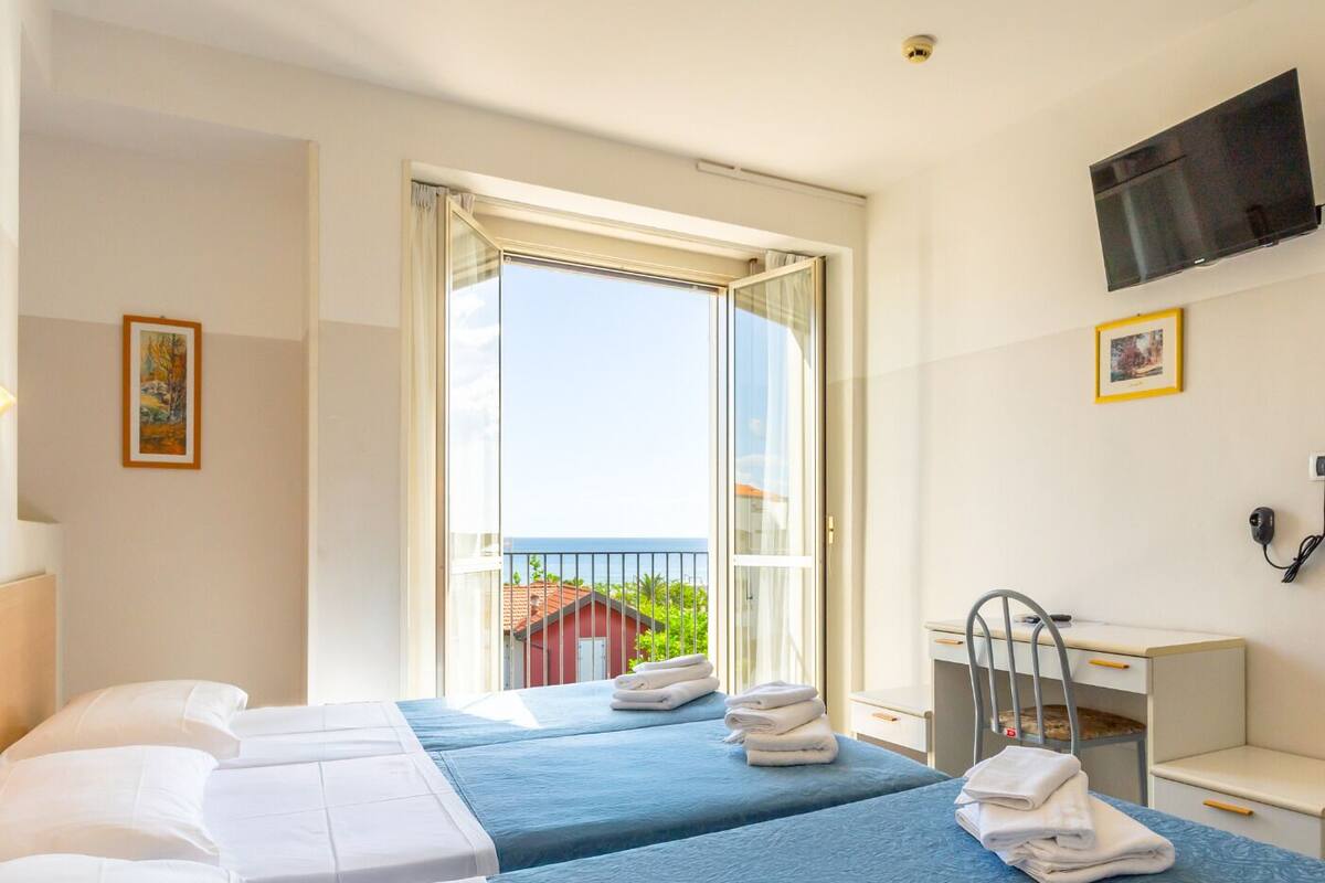 A hotel room with two beds, a desk, a chair, a TV, and a balcony with a view of the sea.