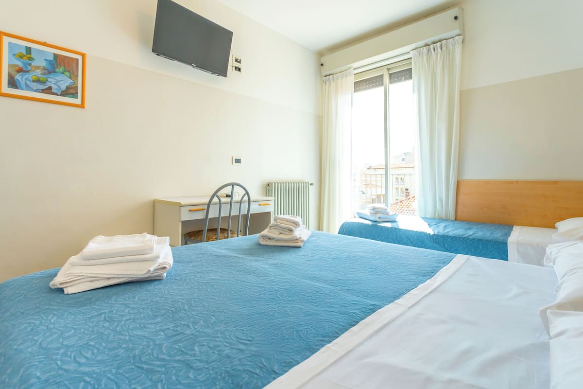 A hotel room with two beds, a TV mounted on the wall, a window with curtains, and a radiator.