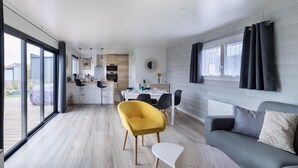 A modern living room with a grey sofa, yellow armchair, and white coffee table. A dining area with a table and chairs is visible in the background. - New Chalet for 8 in a residence with pool (Bretignolles-sur-Mer)