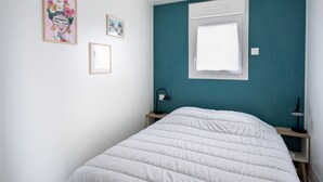 A bedroom with a white bed, teal walls, a window, and two framed pictures on the wall. - New Chalet for 8 in a residence with pool (Bretignolles-sur-Mer)