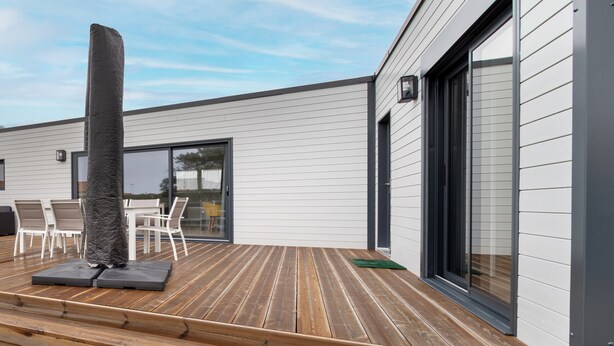 A modern outdoor deck with wooden flooring, a stone pillar, and a glass door leading to a patio area. - New Chalet for 8 in a residence with pool (Bretignolles-sur-Mer)