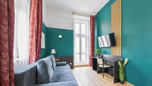 A modern room with a grey sofa, a desk with a chair, a TV mounted on the wall, and a window with curtains. - Hotel Praga 1885 (Prague)
