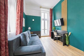 A modern room with a grey sofa, a desk with a chair, a TV mounted on the wall, and a window with curtains. - Hotel Praga 1885 (Prague)