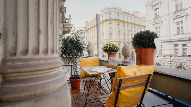 A balcony with a table, chairs, potted plants, and a view of classical architecture. - Hotel Praga 1885 (Prague)