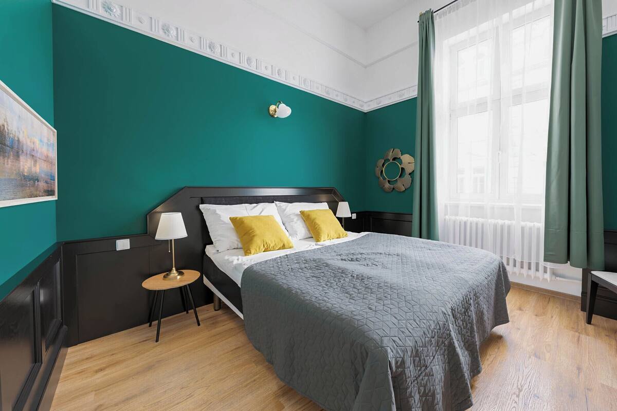 A bedroom with a large bed, teal walls, a white ceiling, a small table with a lamp, and a window with curtains.