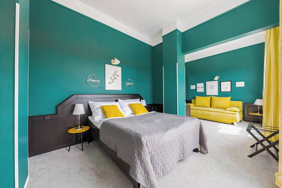 A bedroom with teal walls, a bed with a grey quilt, yellow pillows, and a yellow sofa.