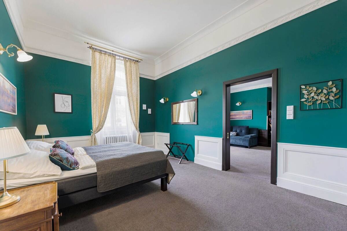A bedroom with a bed, bedside table, lamp, and a view into another room.