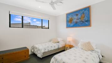 A bedroom with two beds, a ceiling fan, a window with a view of buildings, and a painting on the wall.