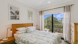 Un dormitorio con una cama grande, una mesita de noche, una lámpara de noche, un aparador y un balcón con vistas a la vegetación y una casa.