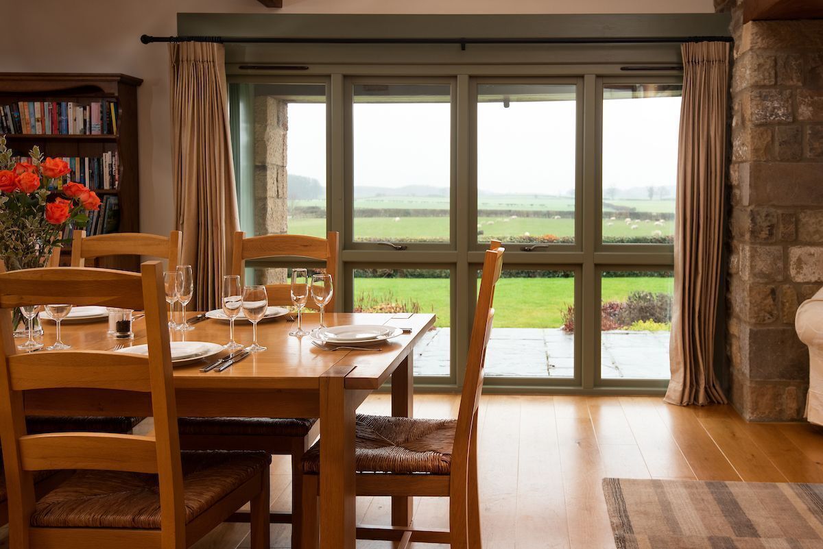 Un coin repas avec une table dressée pour quatre personnes, des chaises en bois, une bibliothèque et une vue sur un paysage verdoyant grâce à de grandes fenêtres.