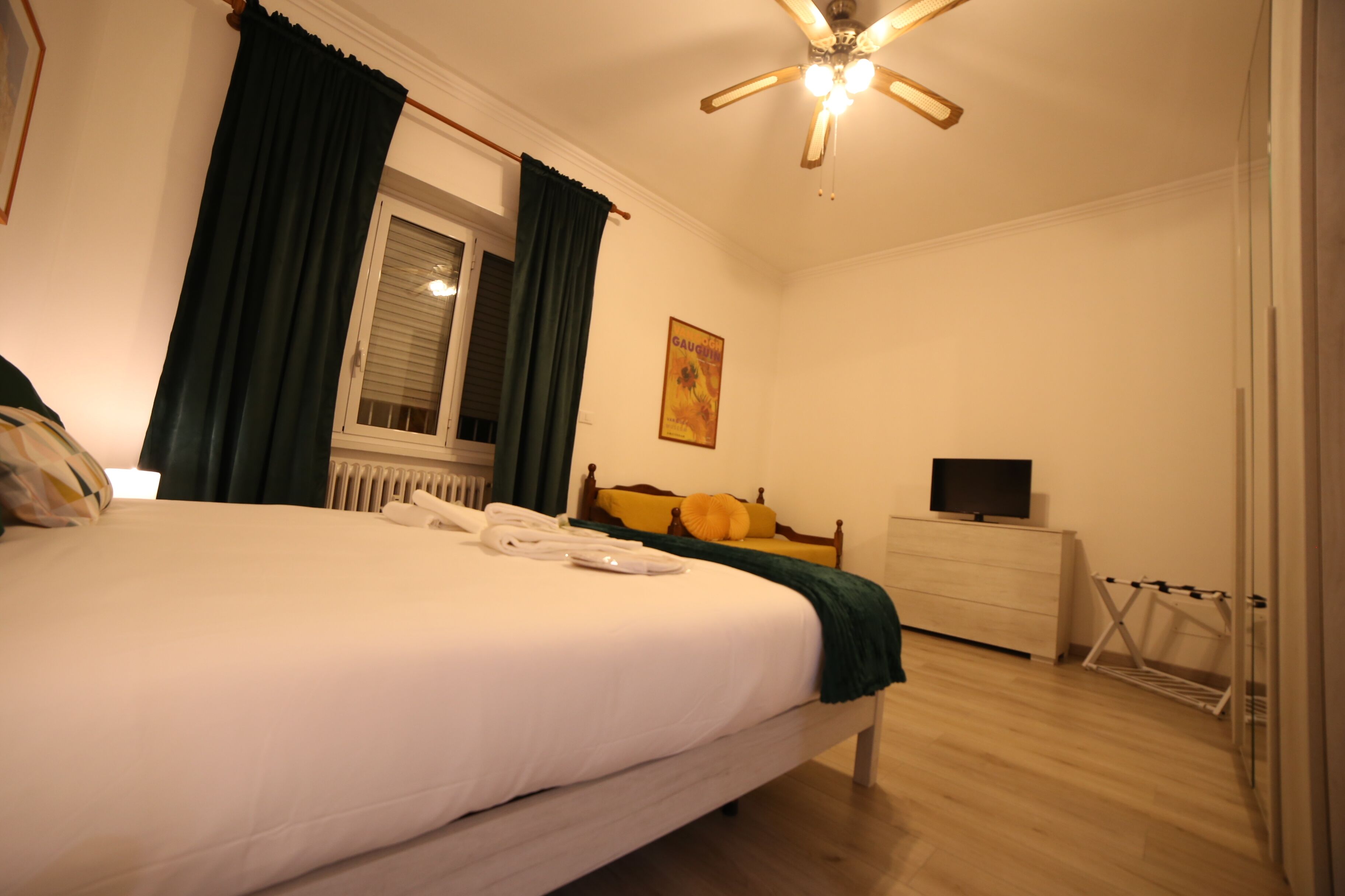 Una cama bien tendida con sábanas blancas, cabecera de madera y manta verde. Se observa una ventana con marco blanco y cortinas verdes, un aparador de madera con un televisor de pantalla plana y un ventilador de techo con luces.