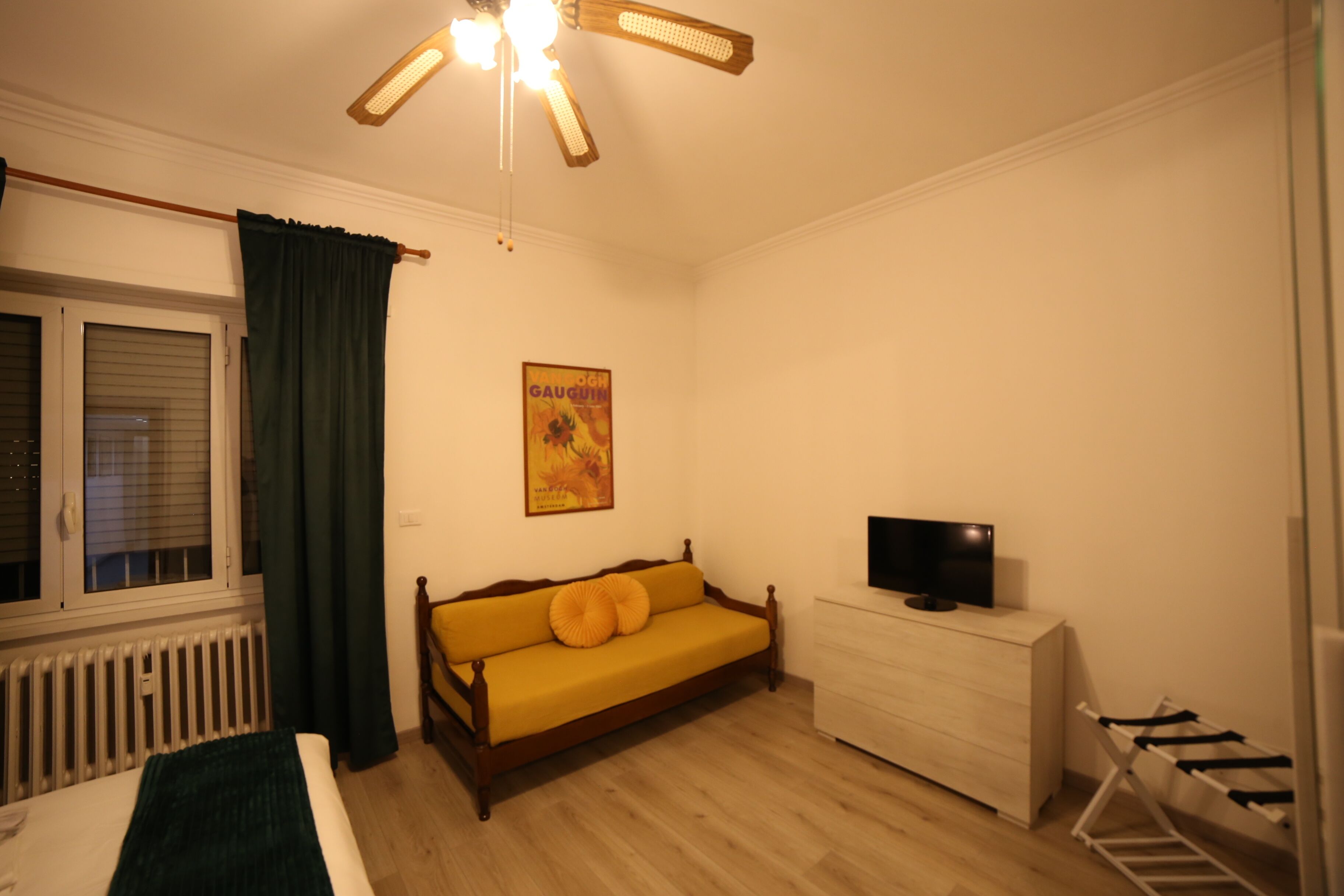 A room with a bed, a wooden sofa, a TV on a dresser, a radiator, and a window with curtains.