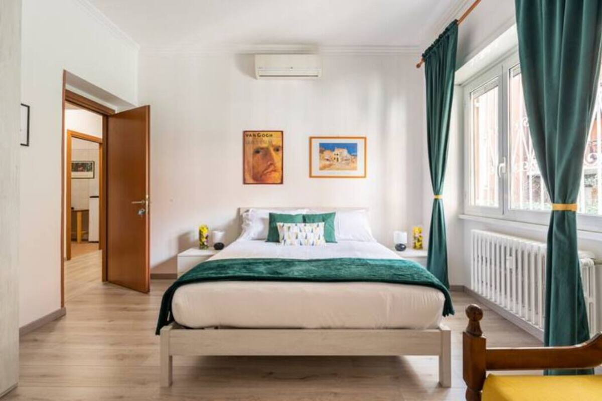 Een slaapkamer met een bed, een nachtkastje, een raam met gordijnen en twee ingelijste foto’s aan de muur.