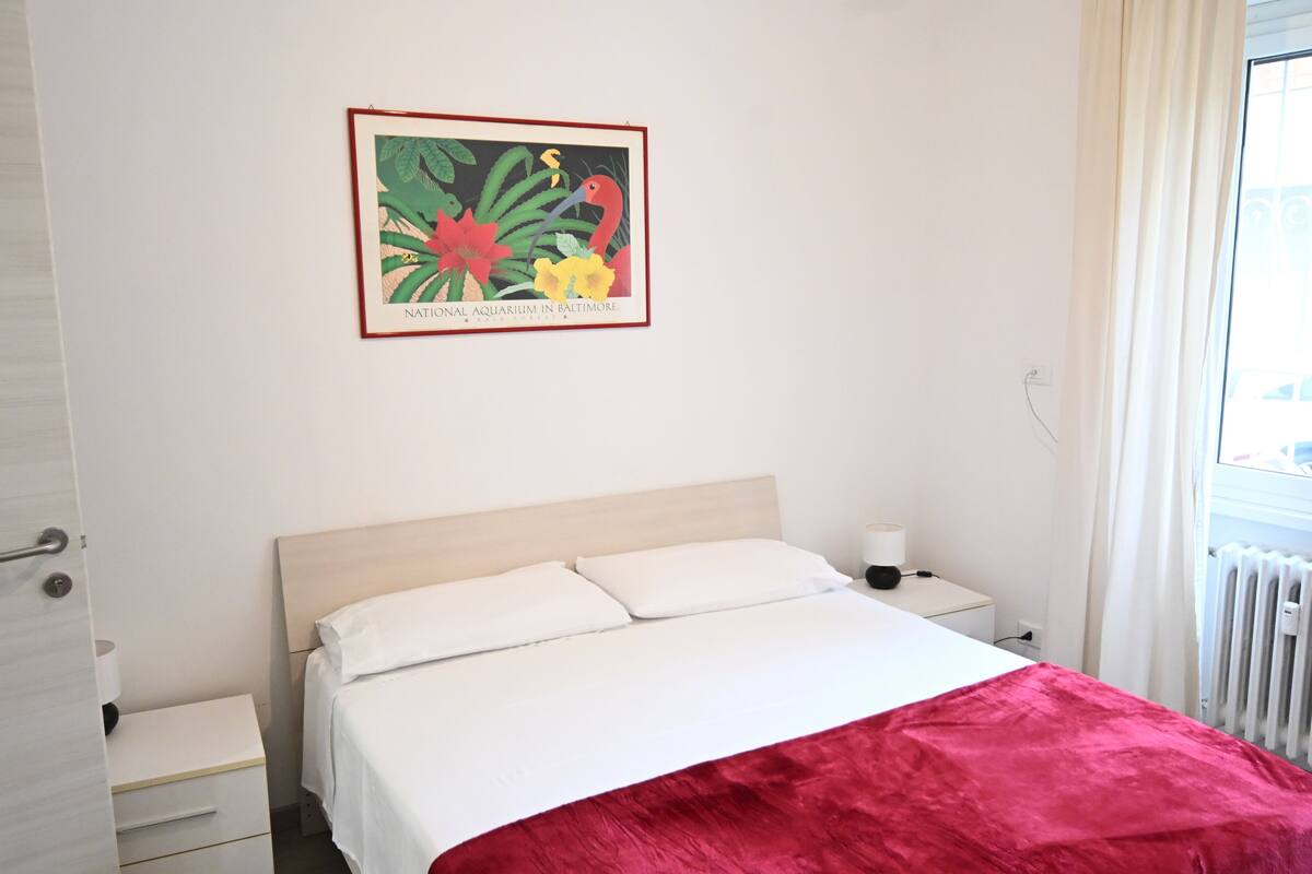 Hotelkamer met een bed, nachtkastje, lamp en een lijst met een afbeelding van een vogel en bloemen aan de muur.