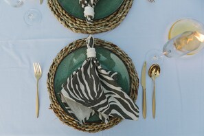 A table set with a woven placemat, a zebra-patterned napkin, a glass wine glass, a gold spoon, and a gold fork.