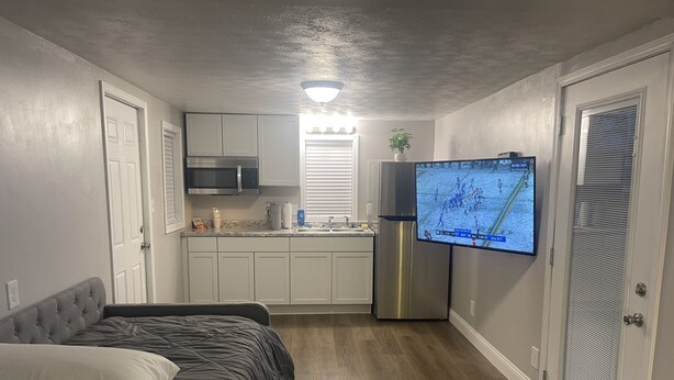 A compact room with a bed, kitchenette, and a TV displaying a sports game.