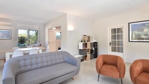 Ett modernt vardagsrum med en grå soffa, två orangefärgade fåtöljer, en matplats med ett vitt bord och stolar samt en bokhylla.