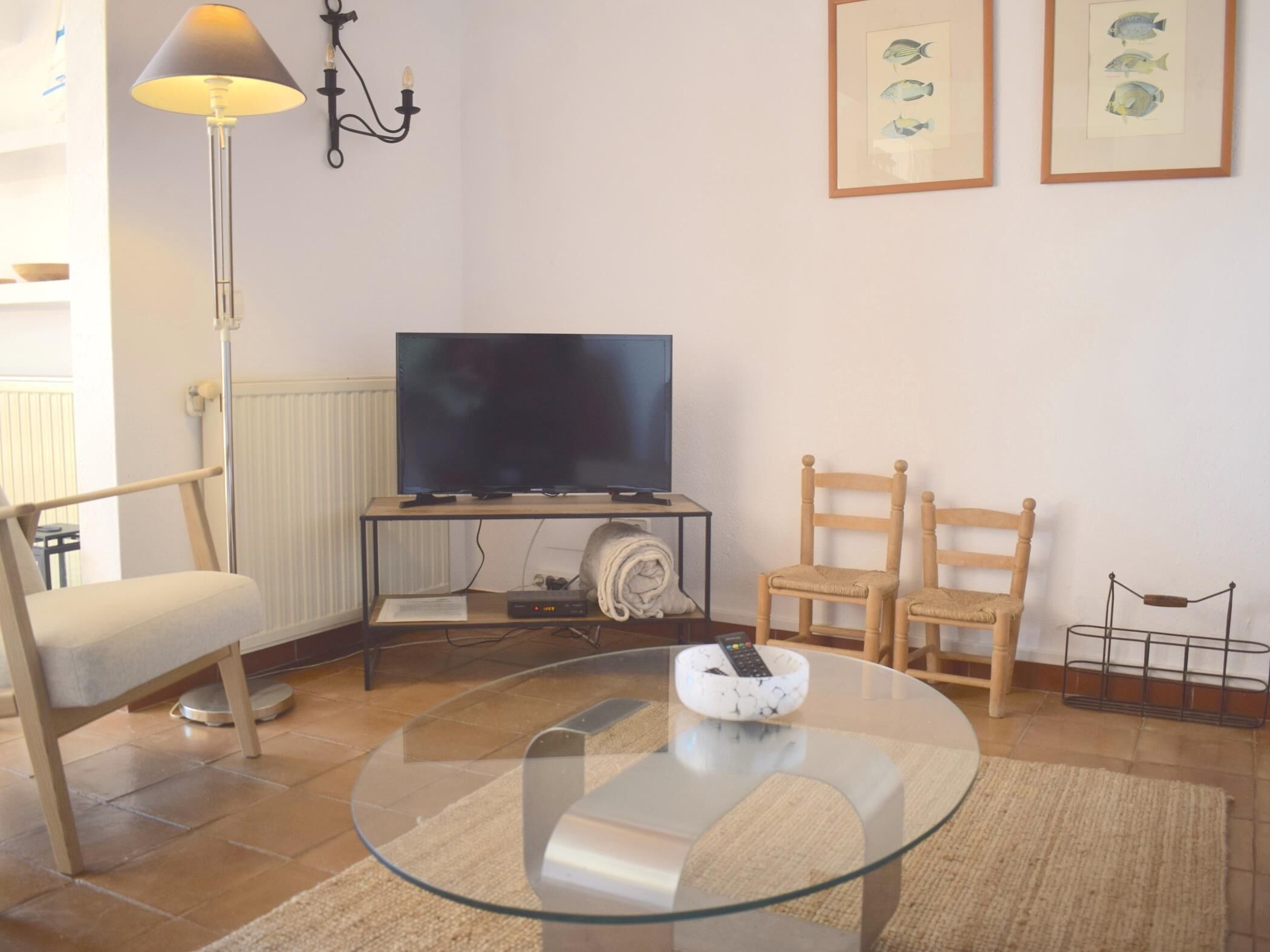 A living room with a television on a stand, a glass coffee table, two wooden chairs, a lamp, and framed fish pictures on the wall.