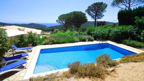 Piscina rectangular con superficie de agua azul cristalina, rodeada por una terraza de piedra y sillas reclinables azules.
