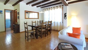 A spacious living room with a dining area, wooden beams on the ceiling, a sofa with orange cushions, and a coffee table with magazines. - Casa Pollo (Begur) (Begur)