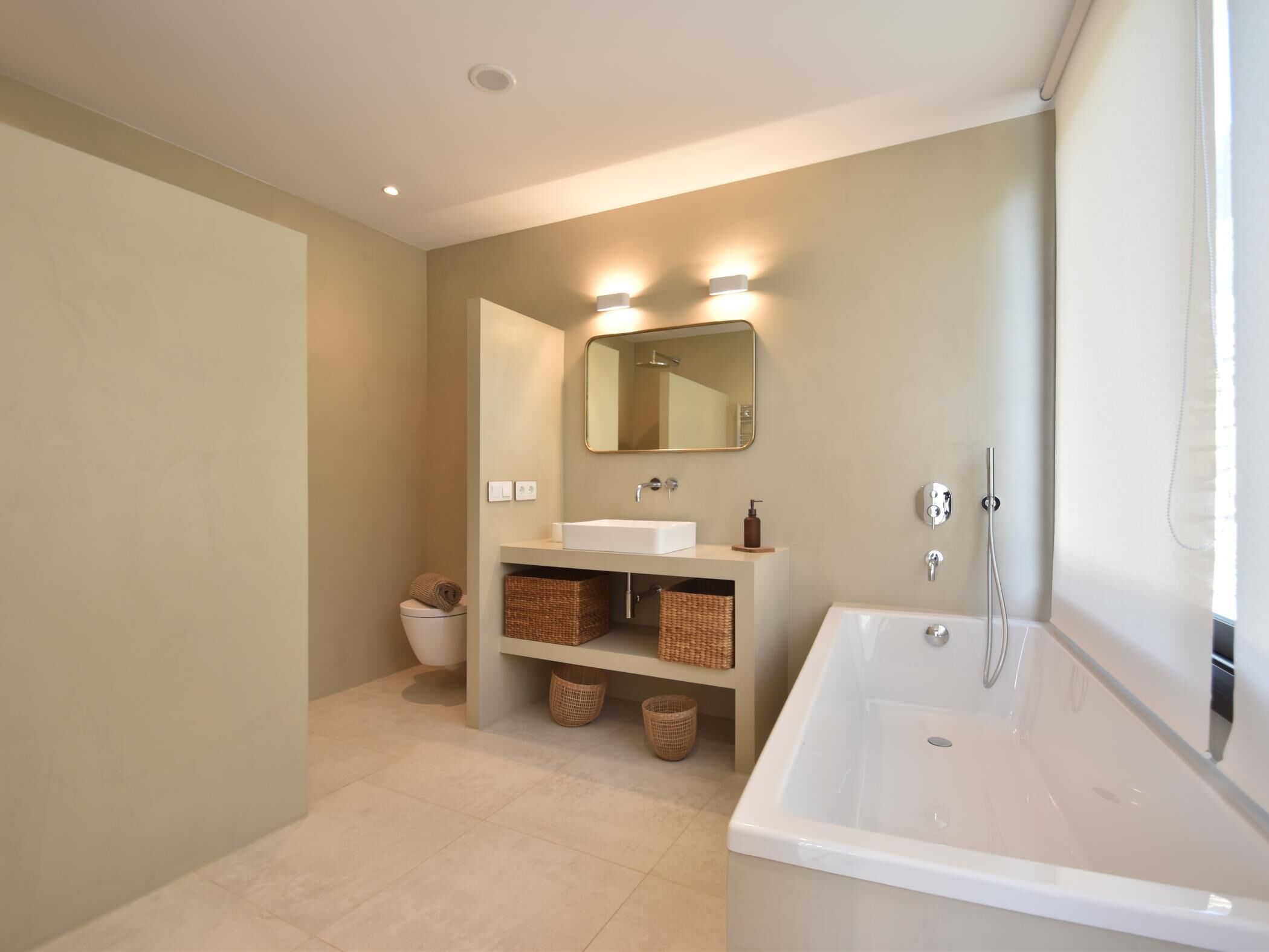 Un baño moderno con una bañera grande, un lavabo y un inodoro. Hay cestas de mimbre debajo del lavabo y un espejo encima.