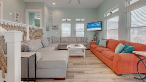 A living room with a grey sectional sofa, an orange sofa, a white coffee table, and a ceiling fan. - The Tipsy Turtle features a playroom for little ones, an arcade game room for bi (Port Aransas)