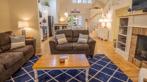 A spacious living room with a brown leather sofa, a wooden coffee table, and a blue patterned rug. A staircase leads to a loft area with a TV and shelves. - Beachwalk Resort Charming Main & Guest House (Michigan City)