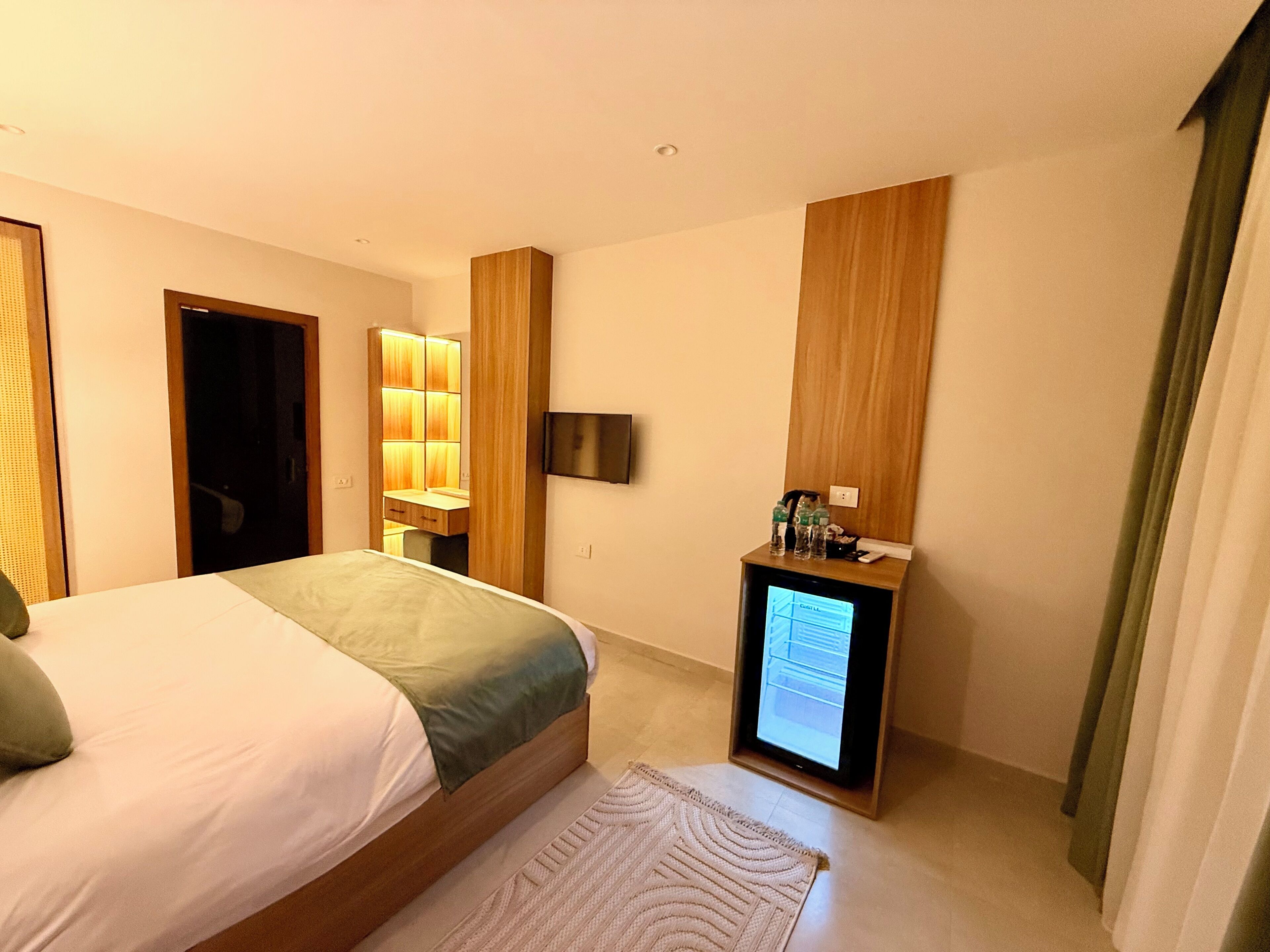 A hotel room with a bed, a desk with a computer, a TV, and a small cabinet with bottles.