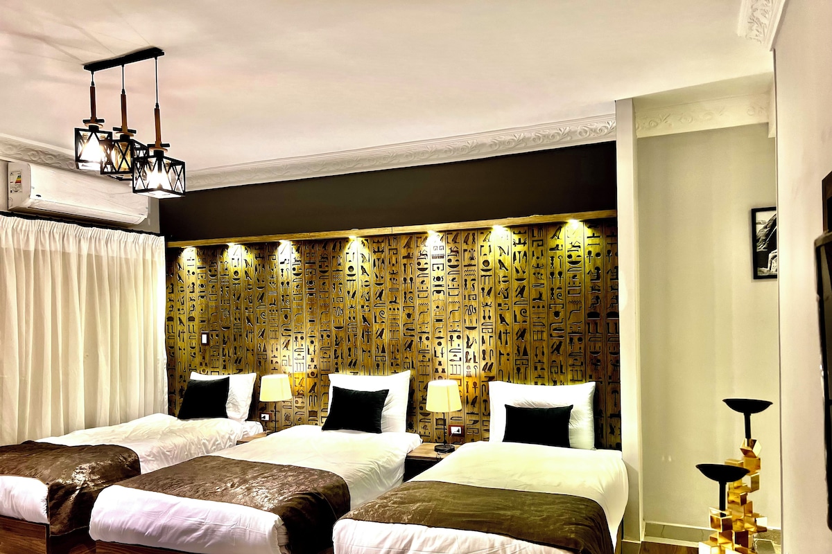 Habitación de hotel con dos camas, un muro decorativo con jeroglíficos, una lámpara de araña moderna y una lámpara de pie.