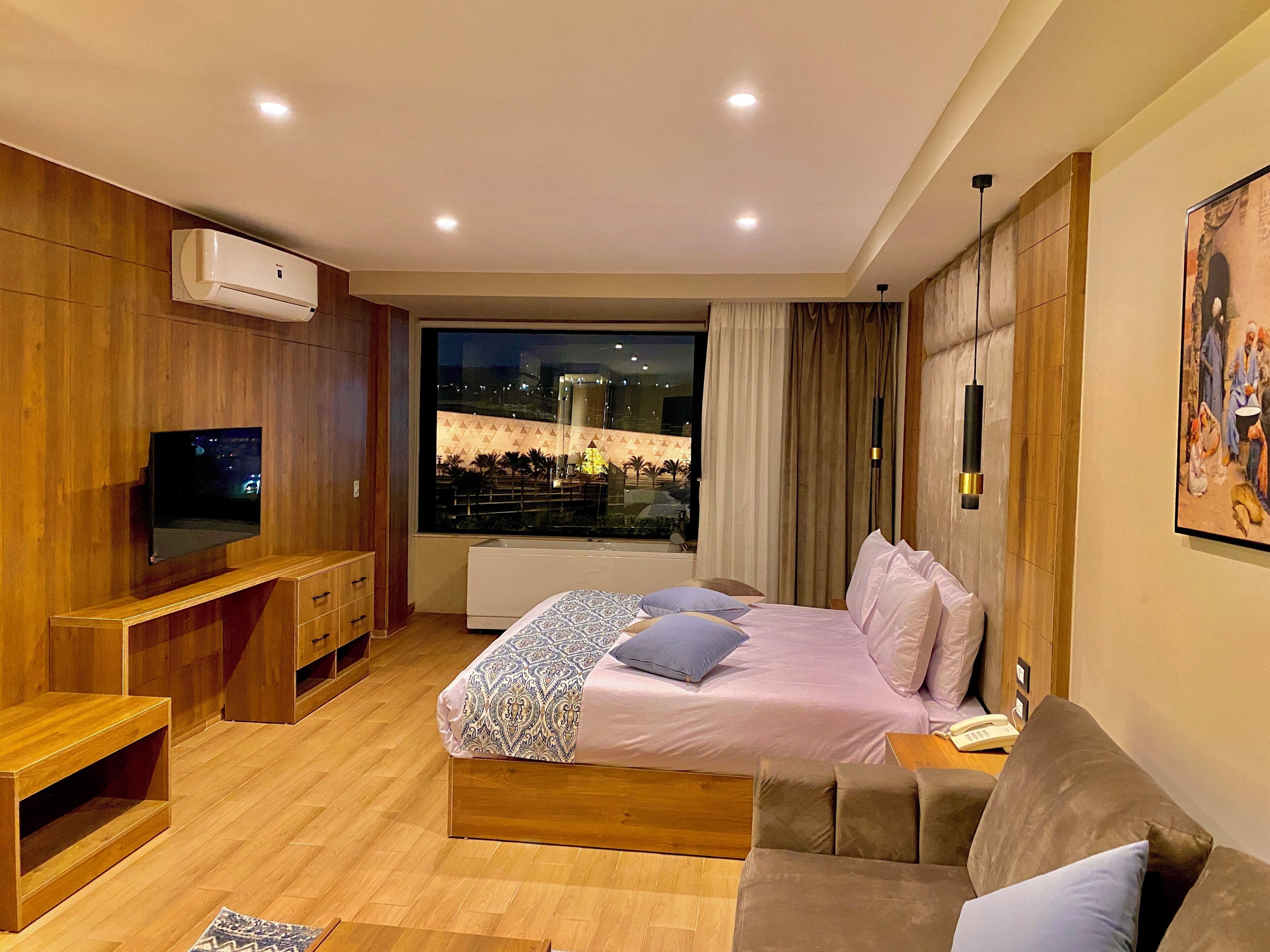 A modern bedroom with a bed, a sofa, a TV, and a large window with a city view.