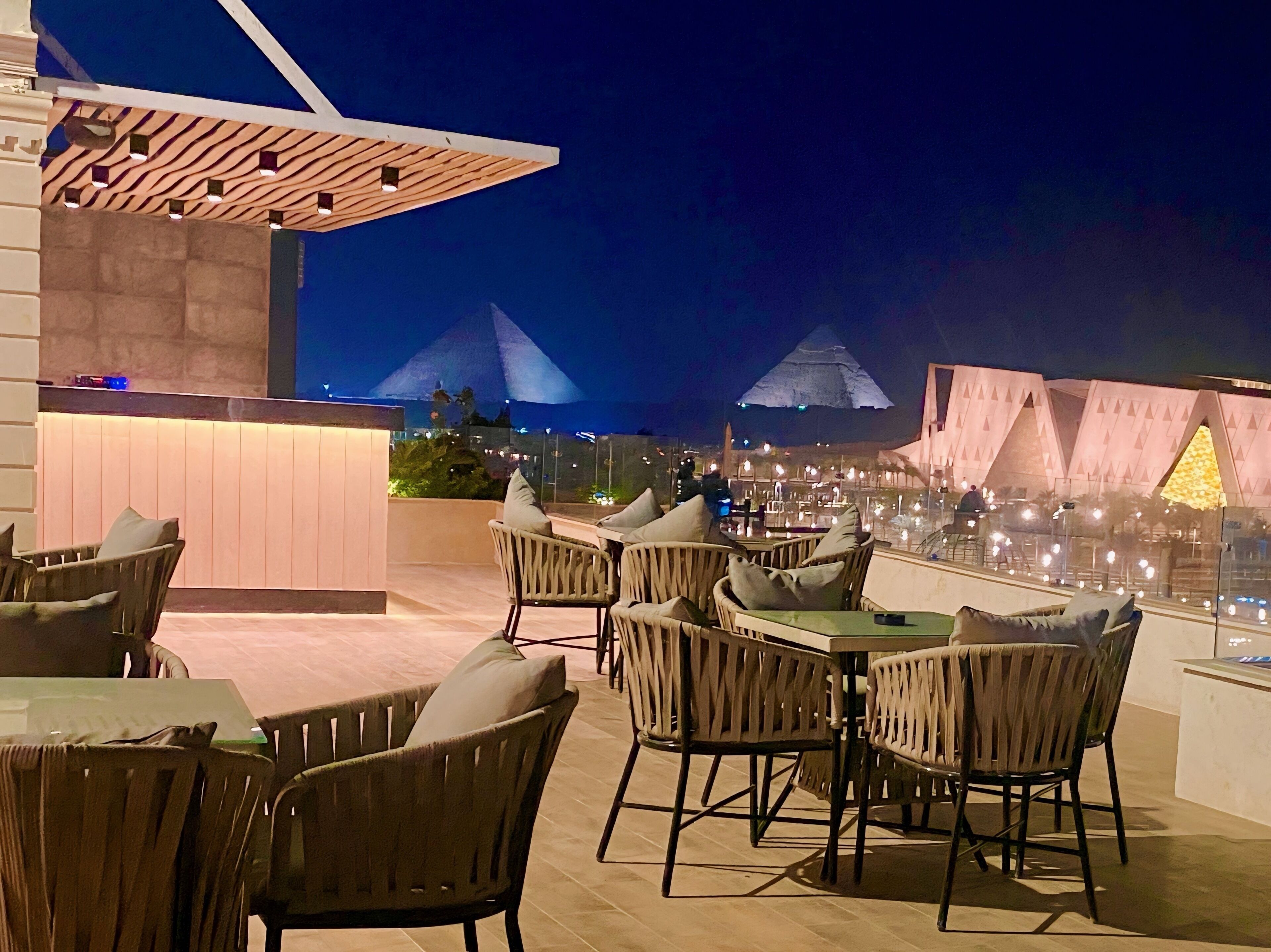 An outdoor dining area with wicker chairs and tables, a modern architectural structure in the background, and a view of a pyramid-shaped building.