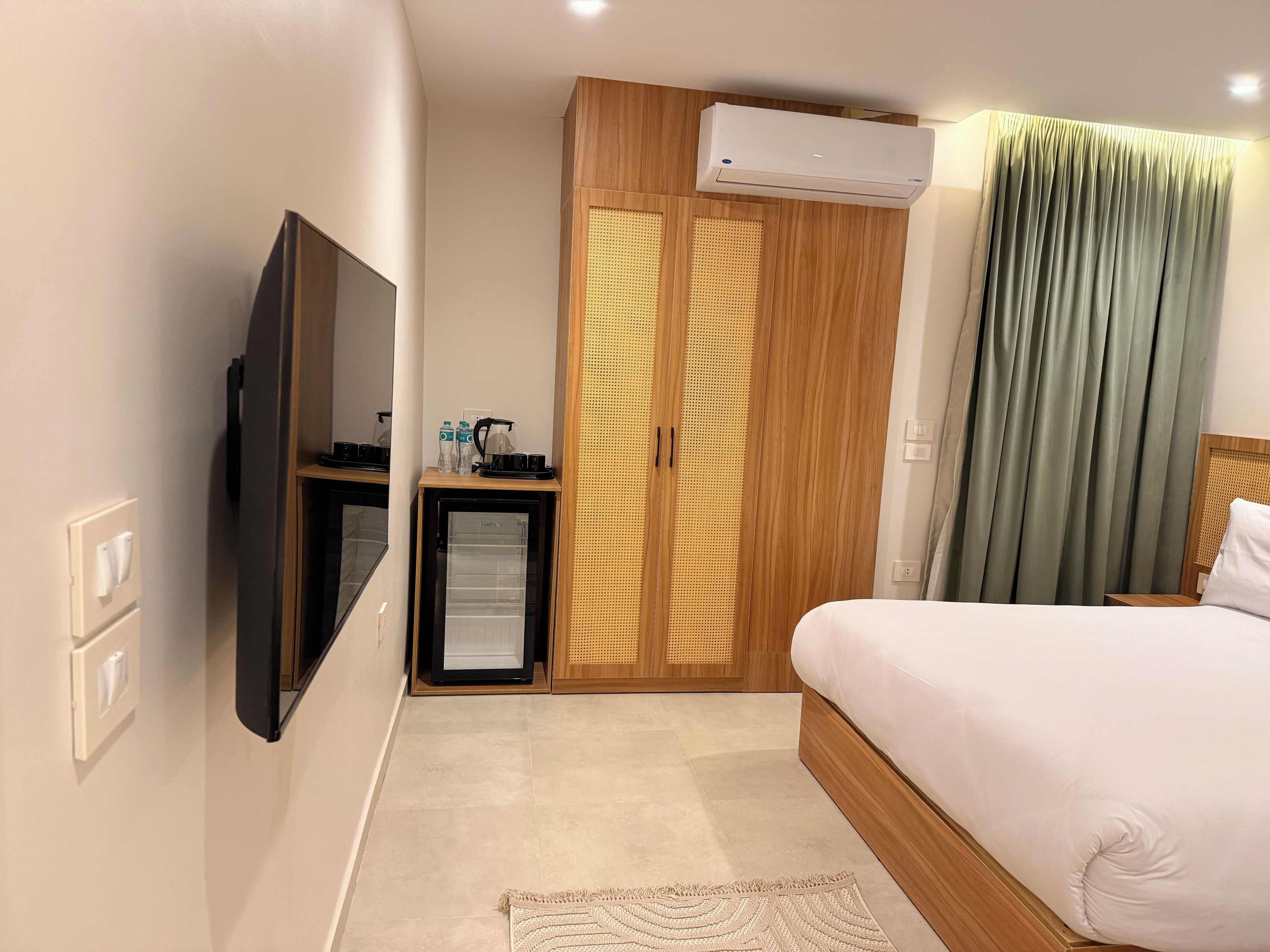 A hotel room with a bed, a TV, a minibar, a wardrobe, and an air conditioning unit.