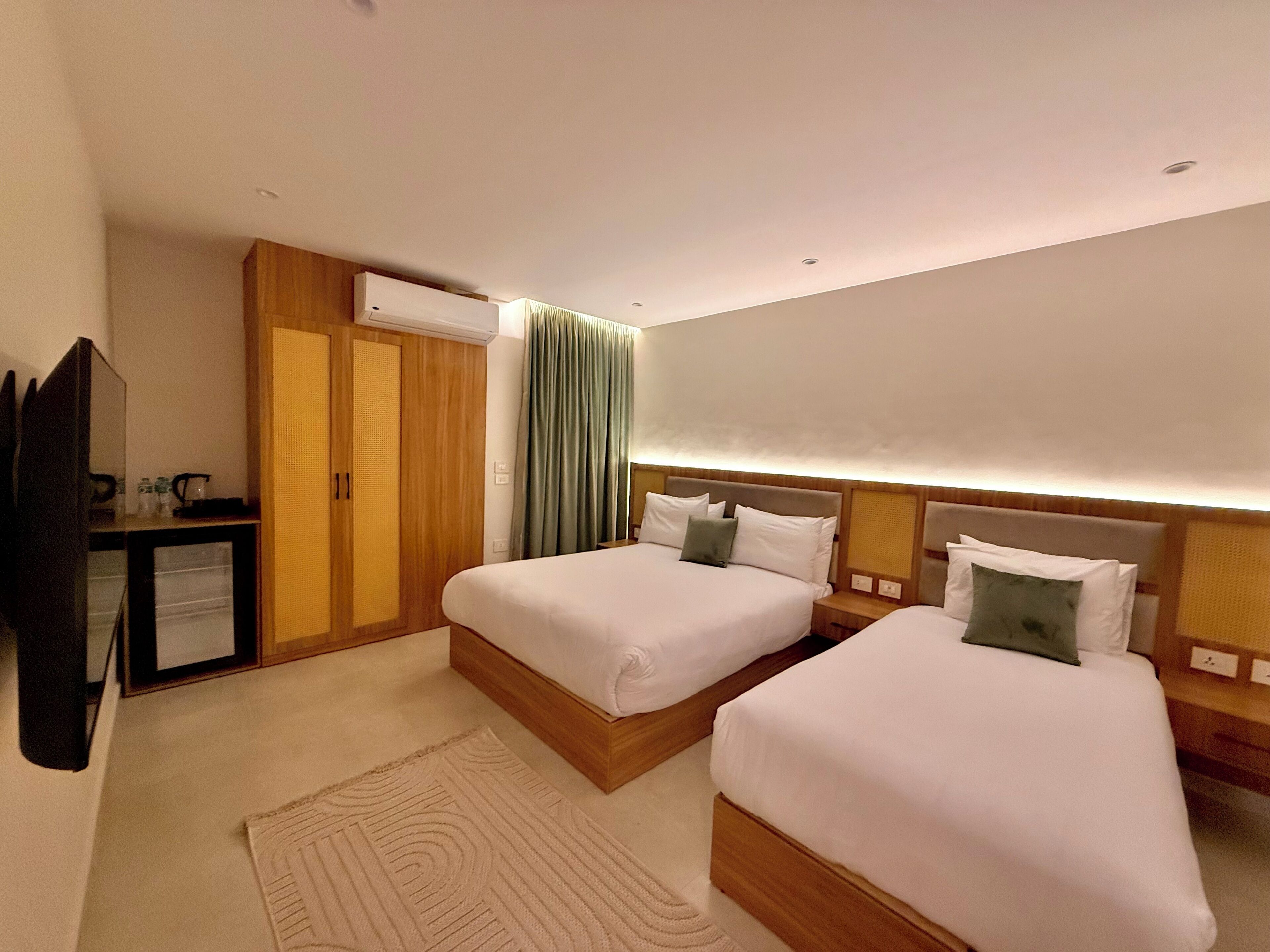 A hotel room with two beds, a wooden wardrobe, a small desk with a lamp, and a large window with curtains.