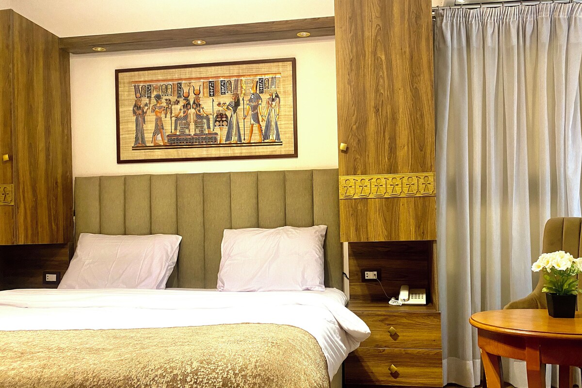 Habitación de hotel con una cama, una mesita con una planta, un teléfono y una obra de arte en la pared que representa figuras del antiguo Egipto.
