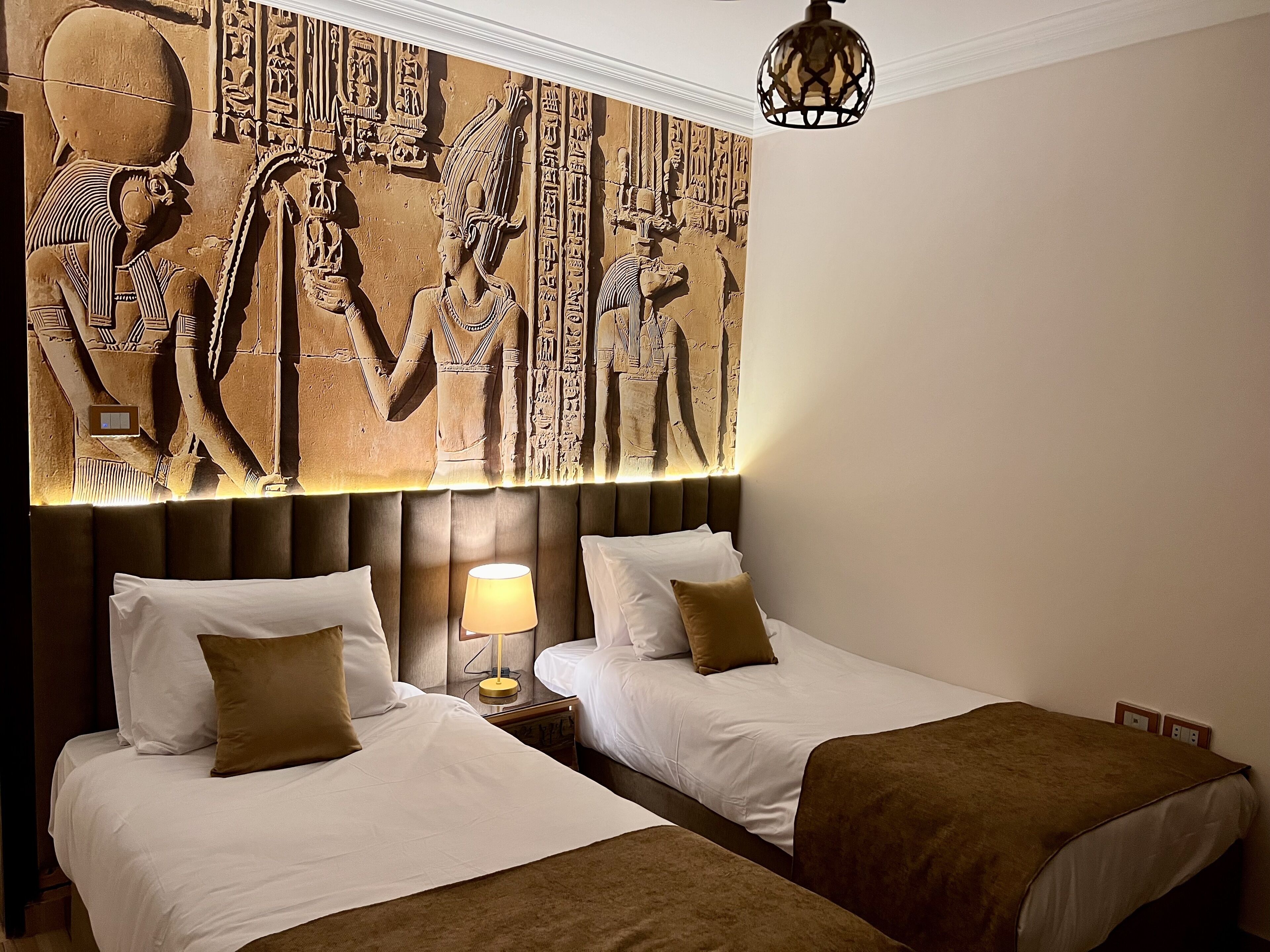 A hotel room with two beds, a wall mural of ancient Egyptian figures, a modern lamp, and a decorative ceiling light.