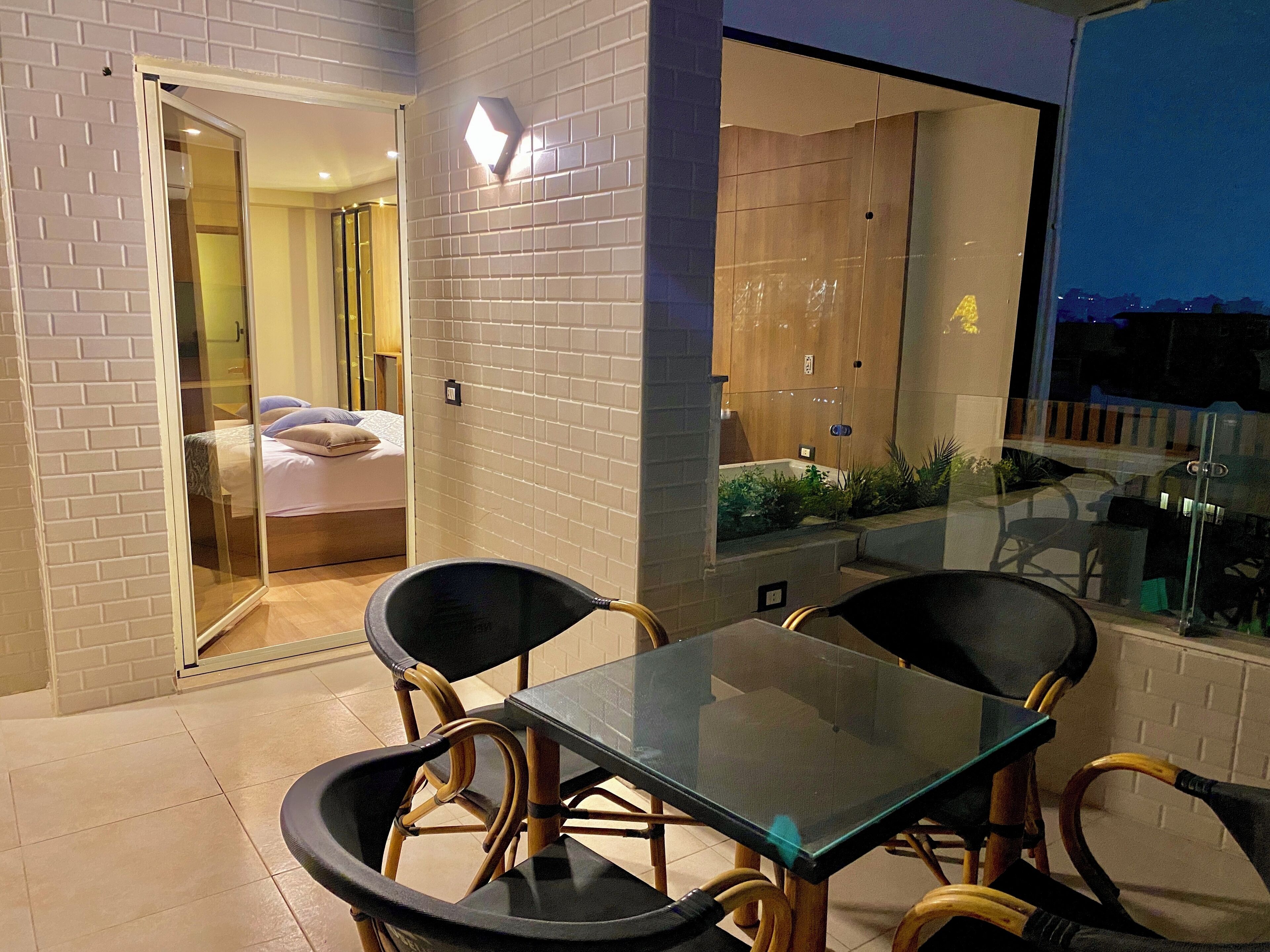 Een balkon met een glazen tafel en stoelen, een schuifpui naar een slaapkamer en een uitzicht op de stad 's nachts.