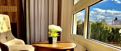 A room with a view of pyramids through a window, a wooden chair, a small table with a vase of flowers, and a curtain.