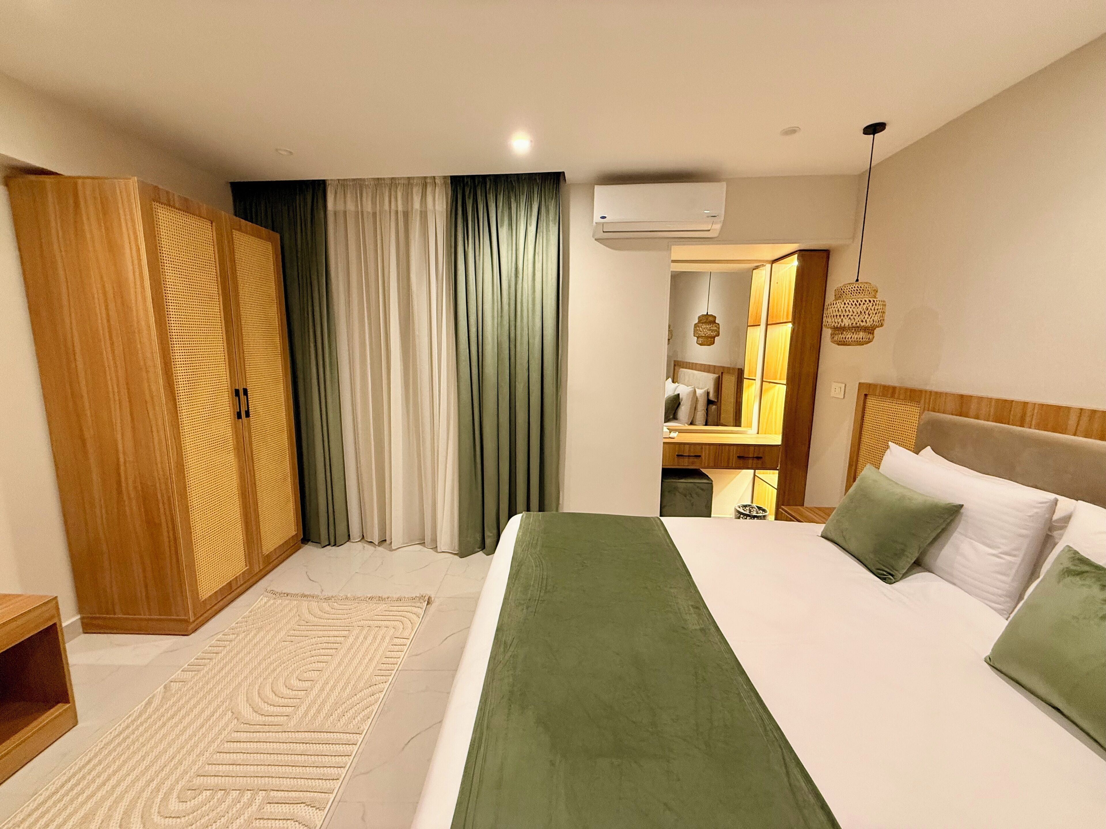 A hotel room with a large bed, a wooden wardrobe, a bathroom visible through a glass door, and a hanging light fixture.