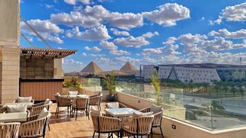 A rooftop terrace with wicker furniture and a glass balustrade overlooking pyramids and a modern building.