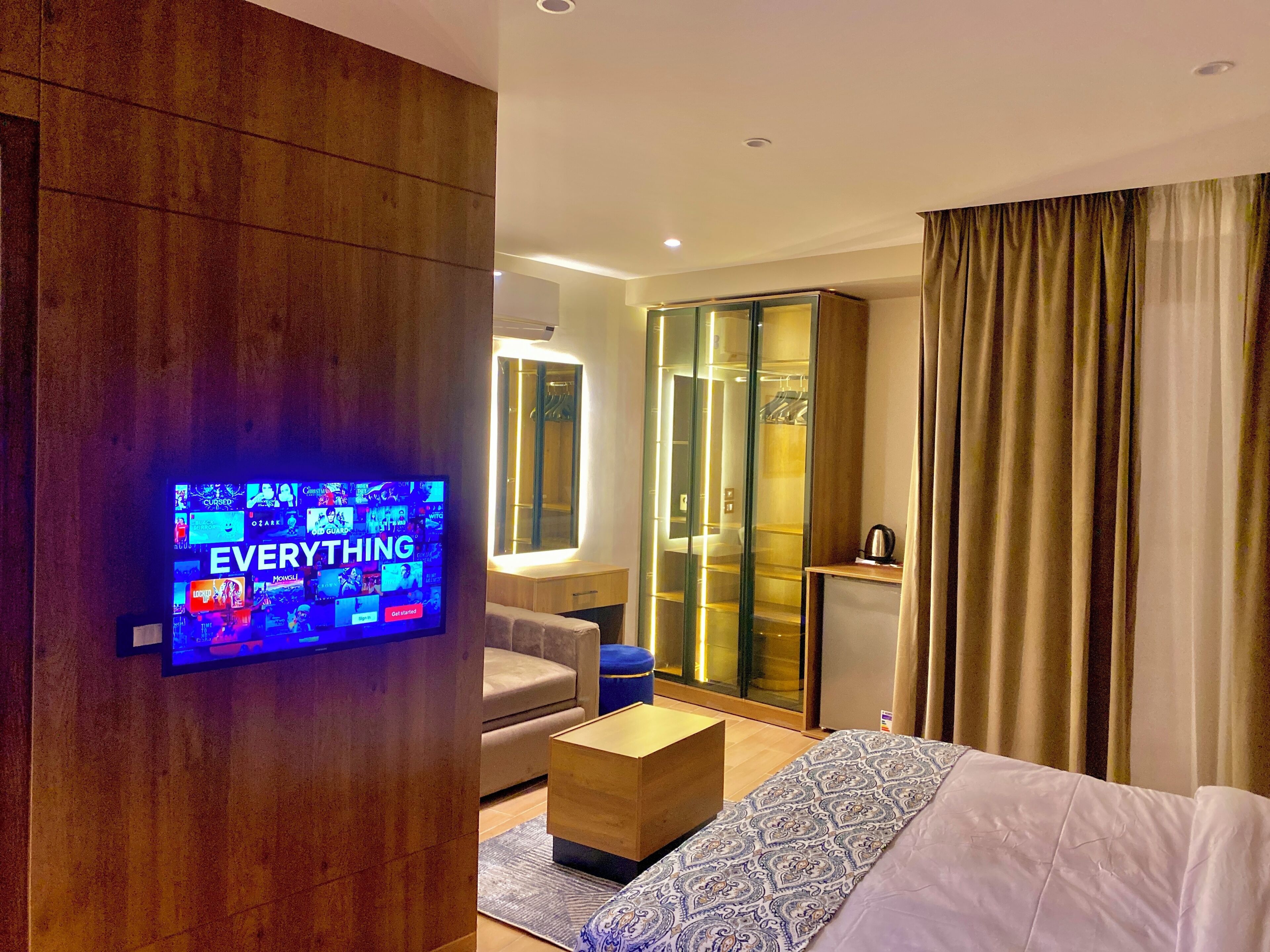 A modern hotel room with a television displaying a movie selection, a sofa, a coffee table, a wardrobe, and a bed.