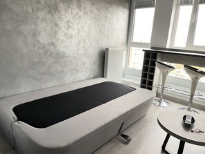 Een moderne kamer met een minimalistisch design, met een enkel bed, een klein rond tafeltje en een raam met jaloezieën.