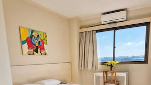 Habitación de hotel con cama, escritorio, unidad de aire acondicionado, ventana con vistas y un cuadro en la pared.