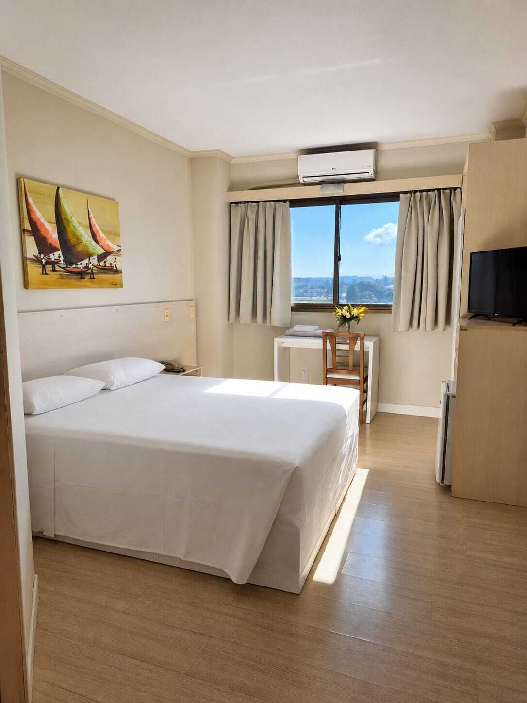 Ein Hotelzimmer mit einem großen Bett, einem Schreibtisch mit Stuhl, einem Fernseher und einem Fenster mit Vorhängen.