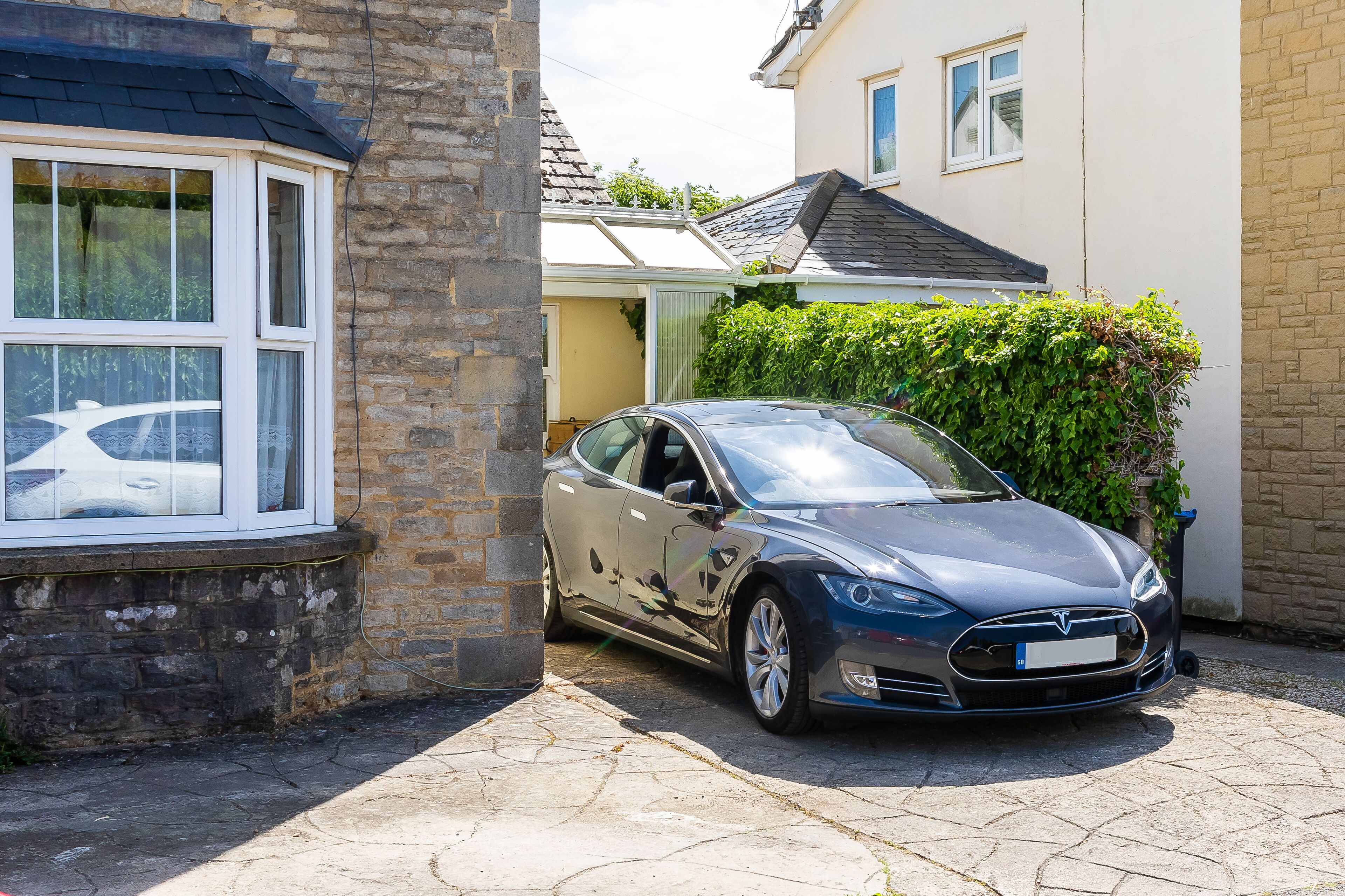 A Tesla Model S parked in a driveway next to a house with a stone wall and a window.