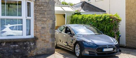 A Tesla Model S parked in a driveway next to a house with a stone wall and a window.