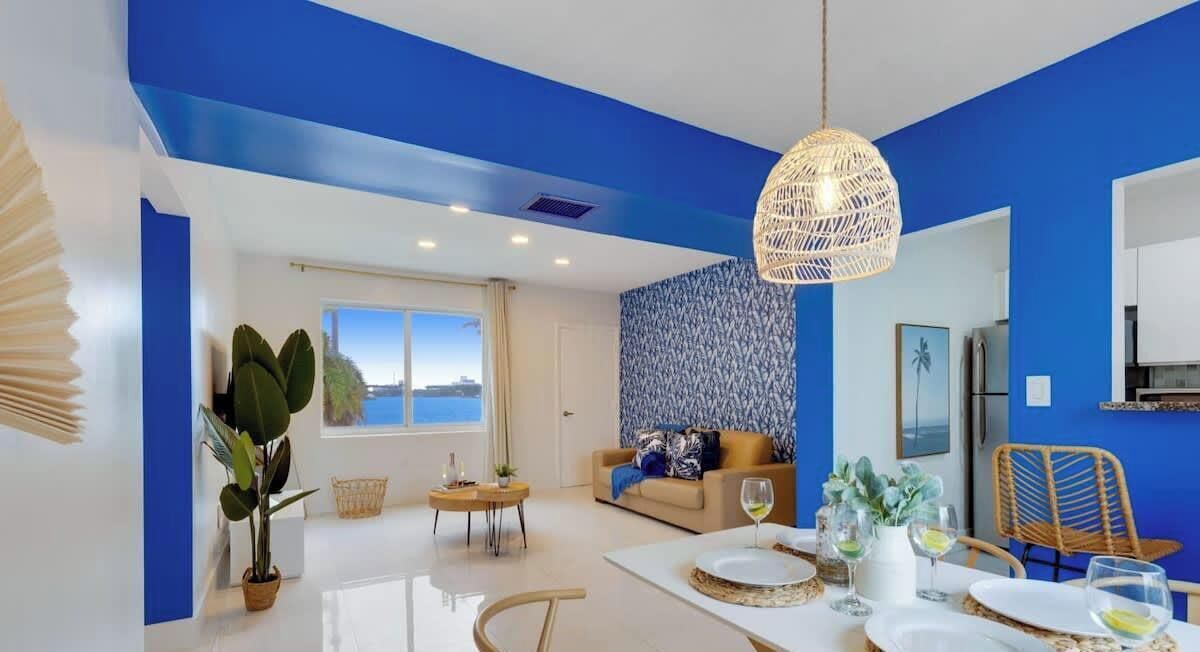 Una sala de estar moderna con una pared azul de acento, un sofá, una mesa de comedor con vasos, y un ventanal grande.